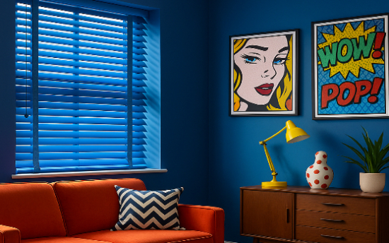 Vibrant pop-art living room with cobalt-blue Venetian blinds over a recessed window, orange sofa with a chevron cushion, mid-century wooden sideboard topped with a yellow desk lamp and polka-dot vase, and colorful comic-style wall art saying “WOW!” and “POP!”.