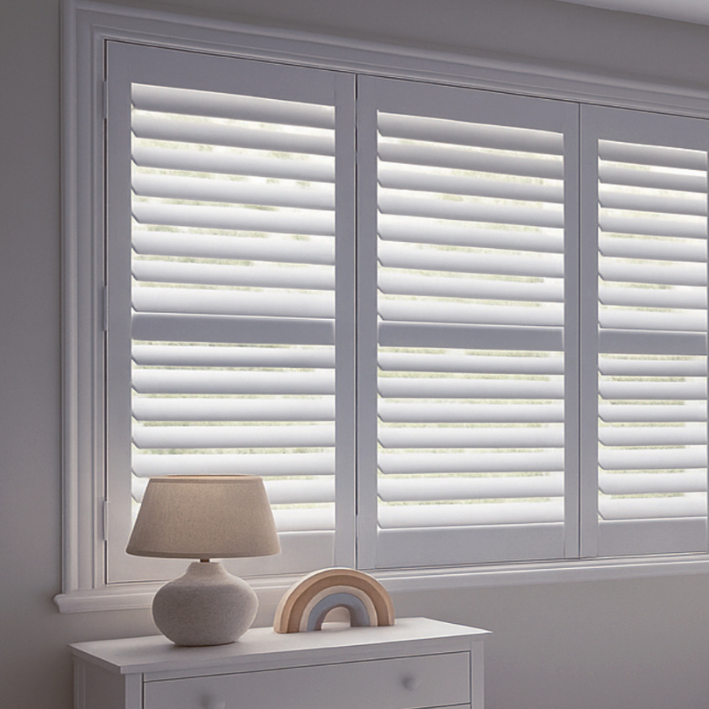 Bright minimalist nursery window with white tier-on-tier plantation shutters, white dresser topped with a stone-textured table lamp and a wooden rainbow toy.