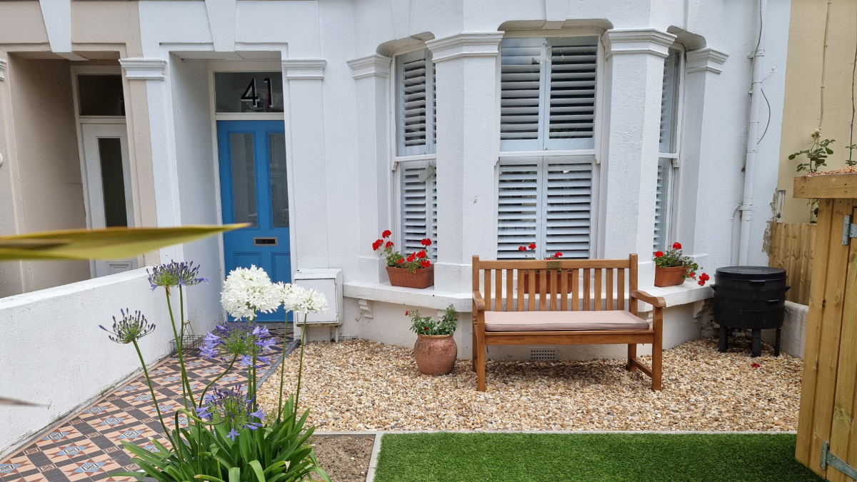 Front garden of a Victorian terrace with white bay-window plantation shutters, vibrant blue front door, patterned tiled path, gravel seating area with wooden bench, and potted flowering plants.