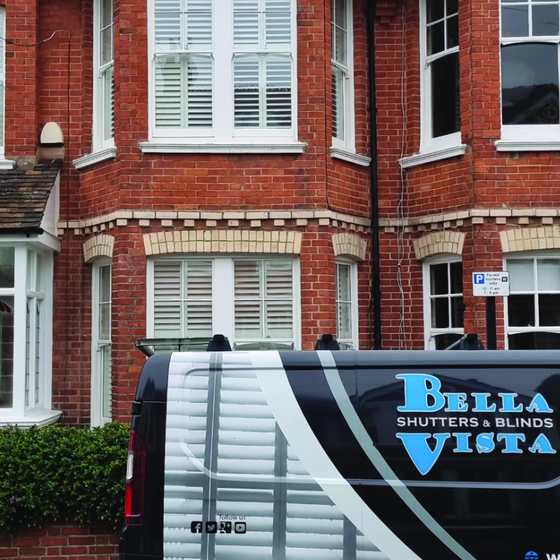 Classic red-brick British townhouse with elegant white plantation shutters installed on the bay windows of both the ground and first floors with Bella Vista Shutters & Blinds van parked outside